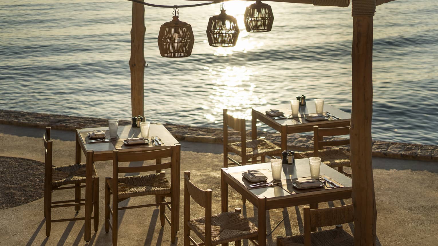 Outdoor taverna next to ocean with three wooden tables and chairs, three wooden lamps