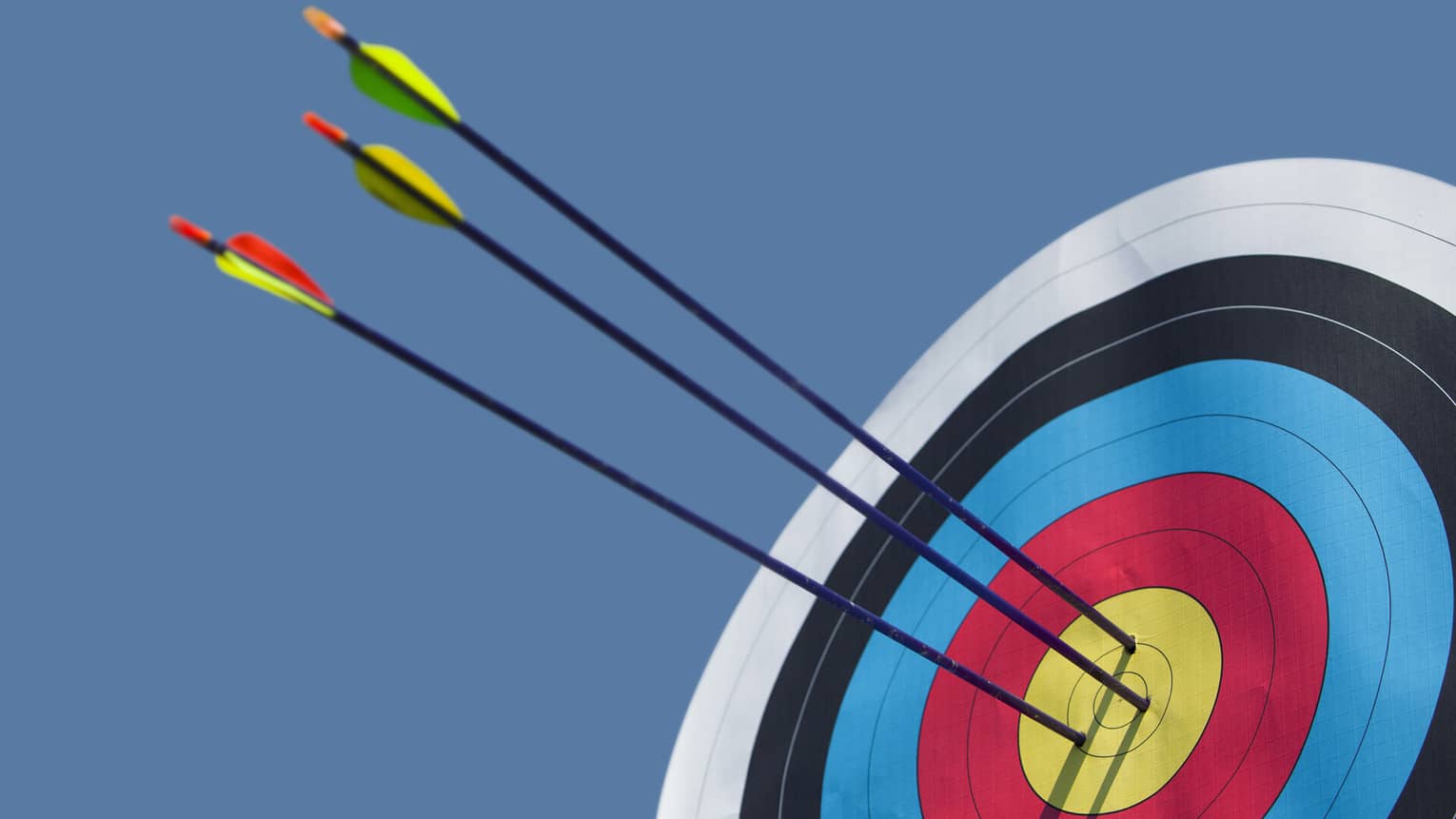 Three arrows with long black shafts pierce the gold circle at the centre of a bull's-eye against the backdrop of a blue sky.