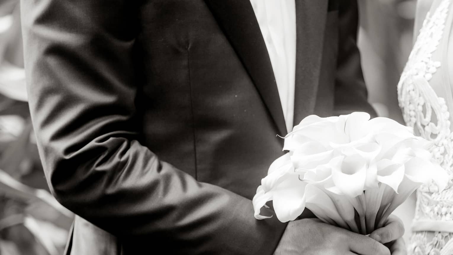 Black and white image of bride wearing lace dress, groom holding wedding bouquet
