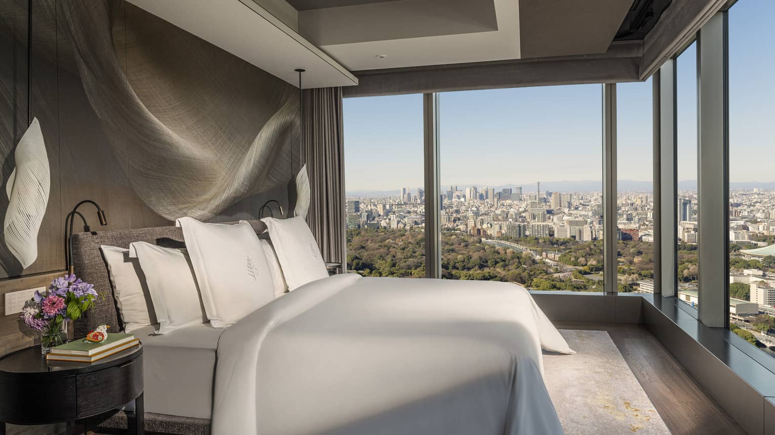A gray upholstered bed, wooden night stand and modern light fixture in a room with stunning city views