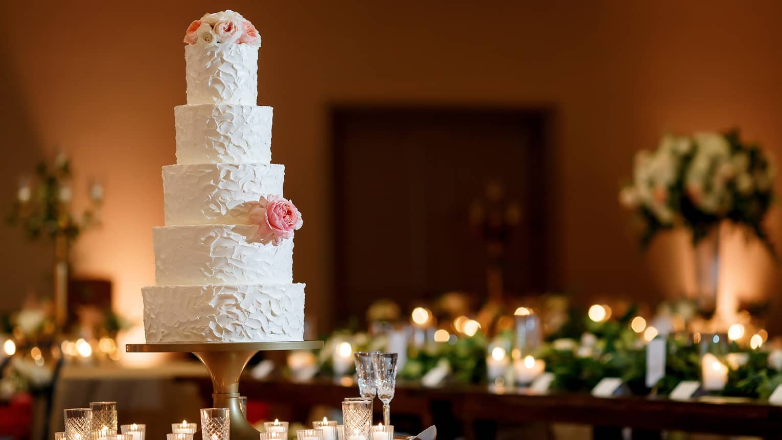 A five-tiered white wedding cake garnished with a white rose and some small white and pink flowers on the top tier , sits on a white round table surrounded by white candles