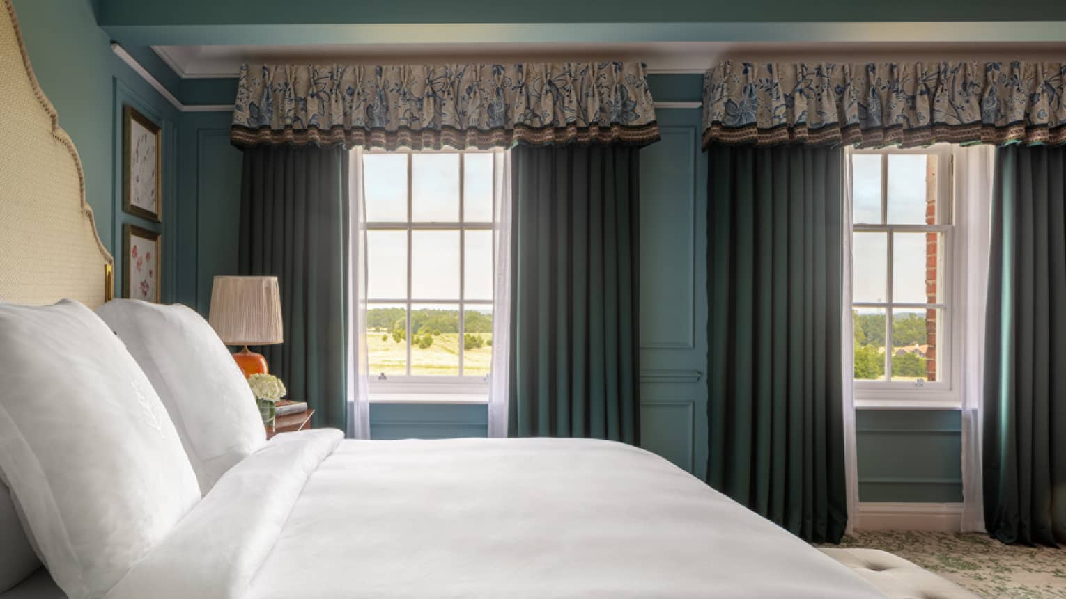 Guest room with bed with white linens, cream headboard and matching bench at foot of bed, twin windows with blue drapes