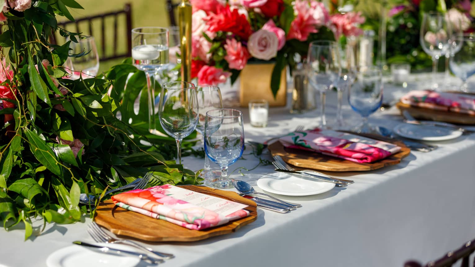 Vibrant table setting with wooden chargers, floral napkins, glassware, pink floral arrangements