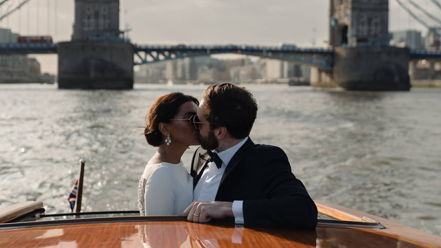 A bride and groom kissing on a boat with a large bridge behind them.