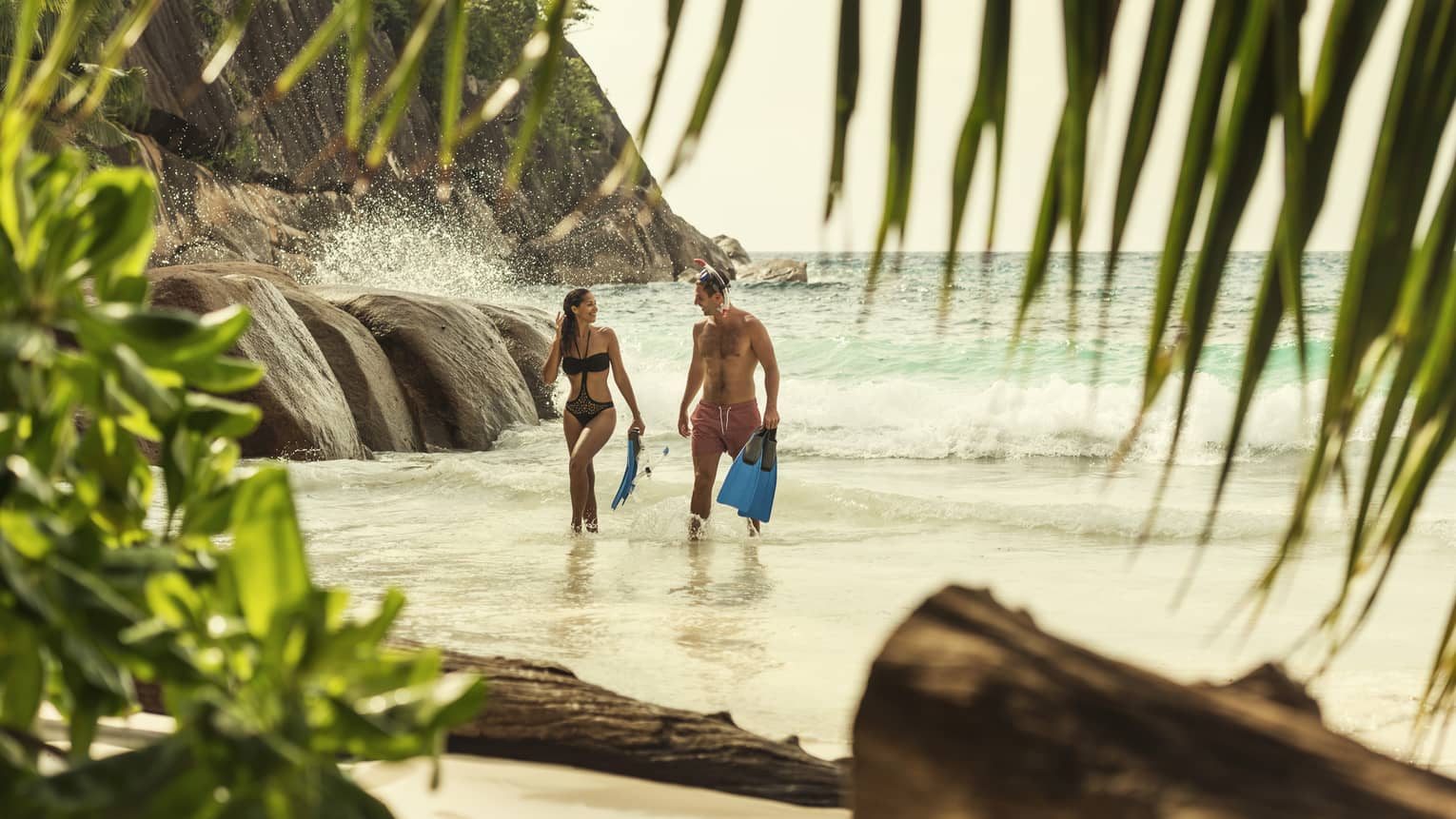 Man and woman in swimsuits hold snorkelling fins, walk from the ocean to beach