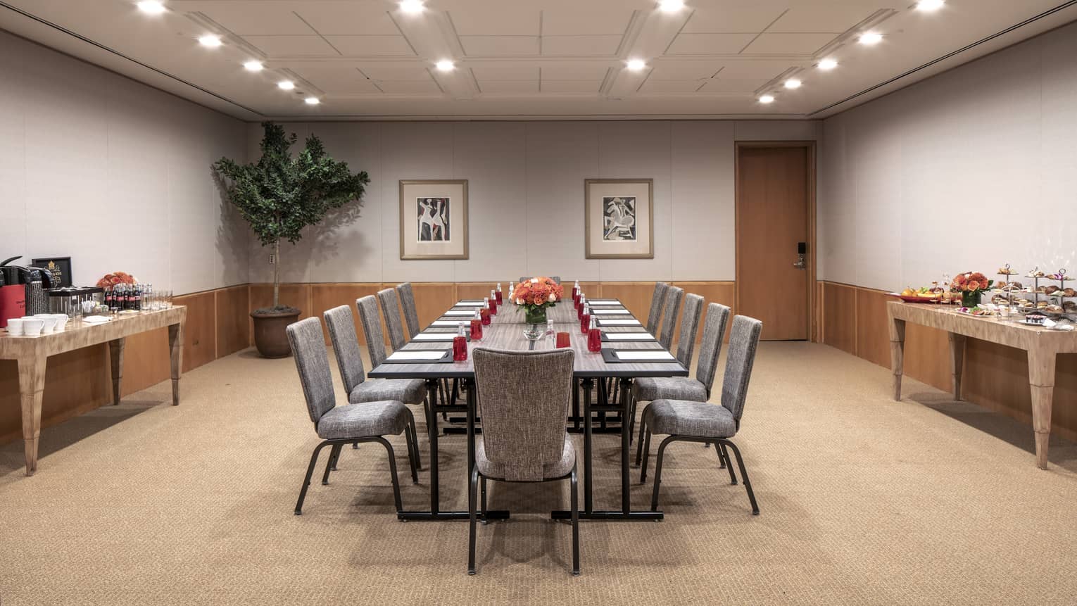 A boardroom with a long rectangular gray table that is set with red water glasses, a black folder, white paper and a orange floral centerpiece. 