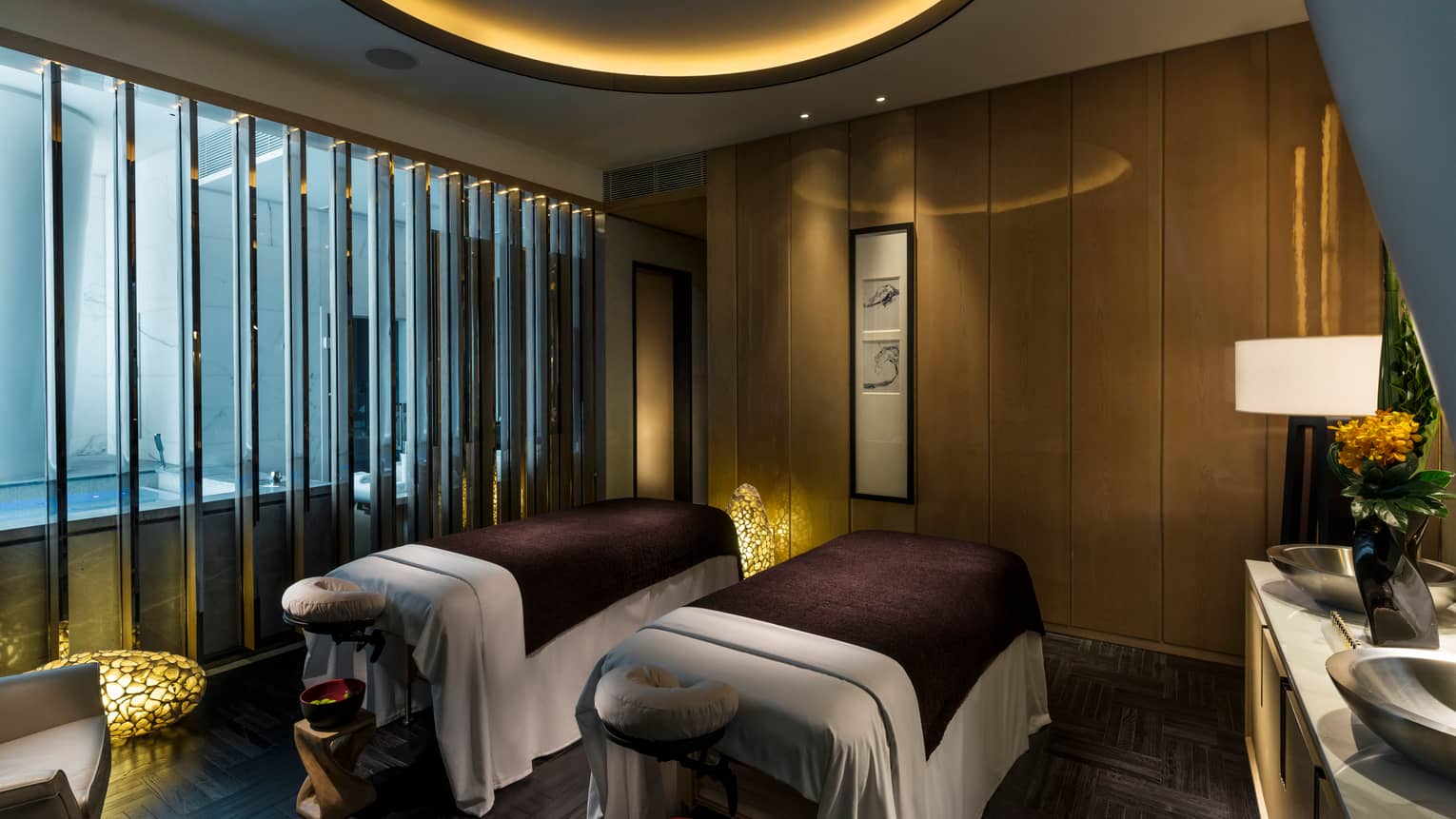 Two massage tables with burgundy blankets side-by-side in dark room with dim lights, lanterns, near tub  
