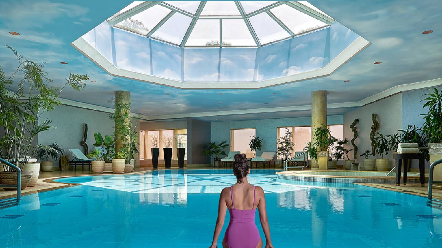 Woman in swimsuit seated on edge of indoor pool with blue ceiling and skylight above