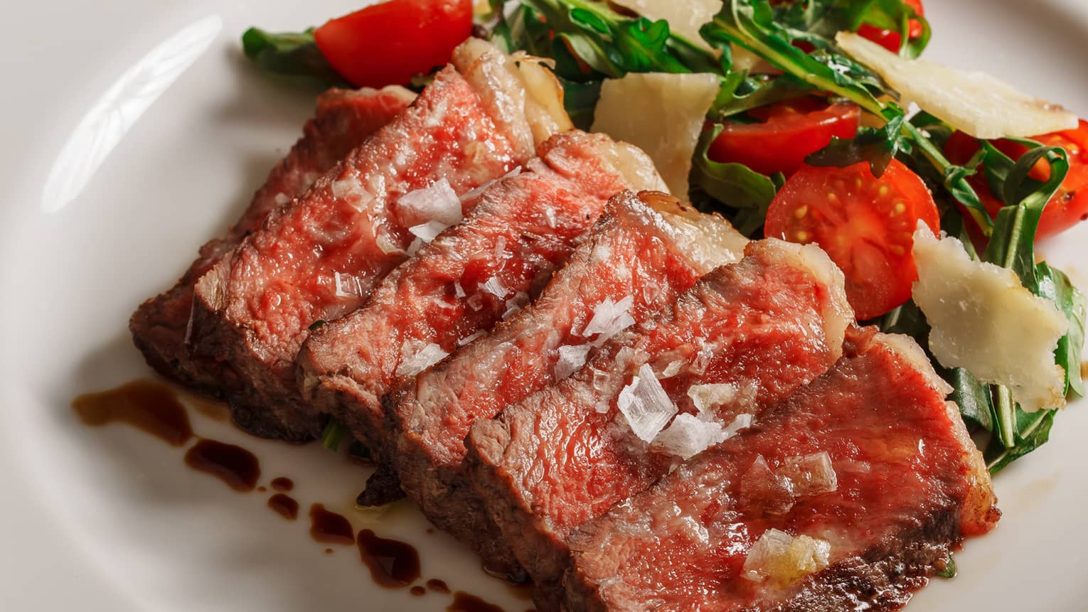 Medium rare grilled beef slices with tomato, rocket and parmesan side salad on white plate