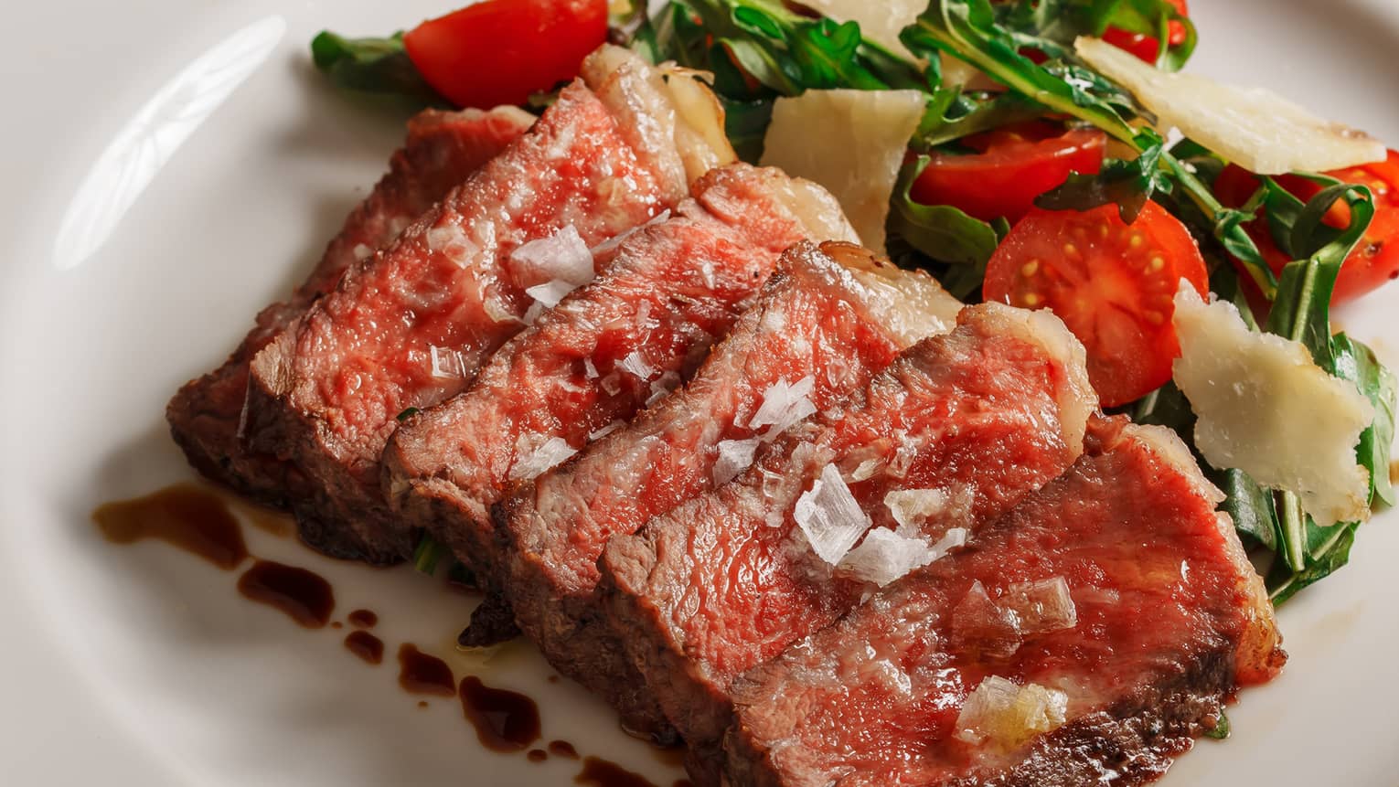 Medium rare grilled beef slices with tomato, rocket and parmesan side salad on white plate