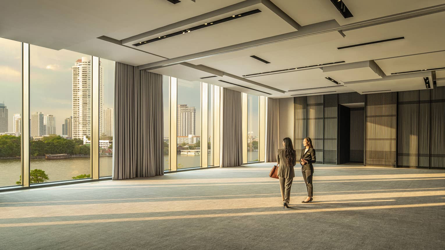 Two woman stand in the empty Conservatory Ballroom gazing out the floor-to-ceiling windows at the Bangkok skyline 