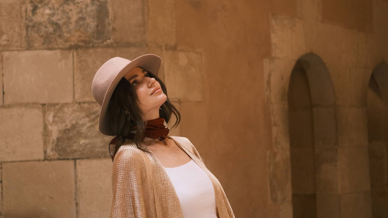 A modestly dressed guest reclines on a stone bench gazing upward, behind them stone walls and arched doorways.