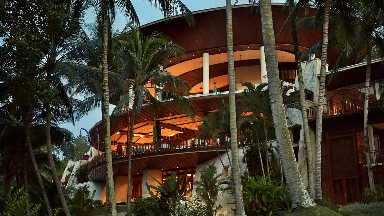 Four-storey curved balconies around resort building at dusk, lit with orange lights, surrounded by palms