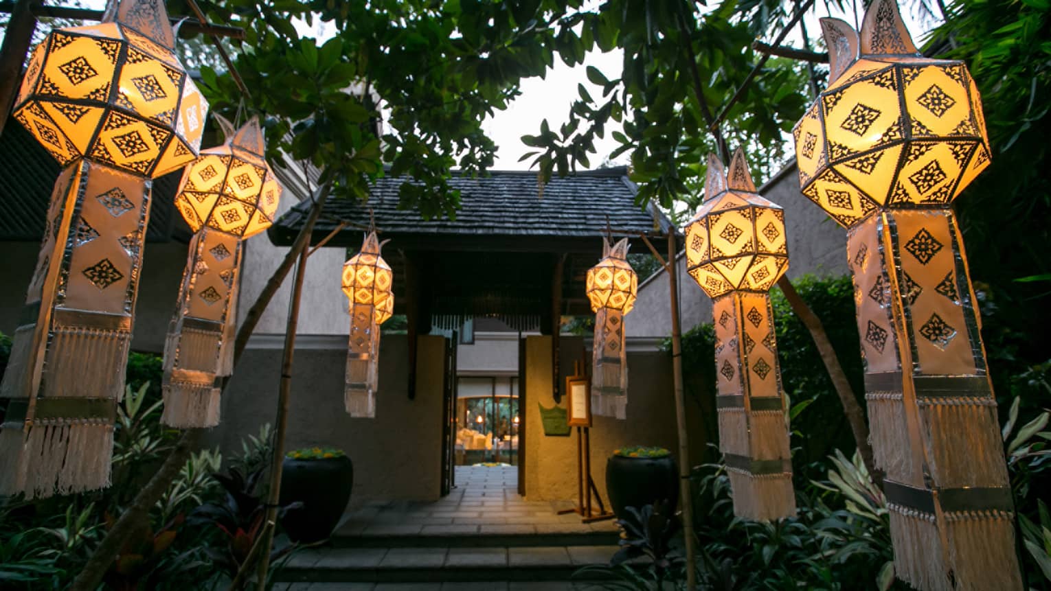 Long glowing Thai lanterns with fringes hang from trees along pavilion walkway