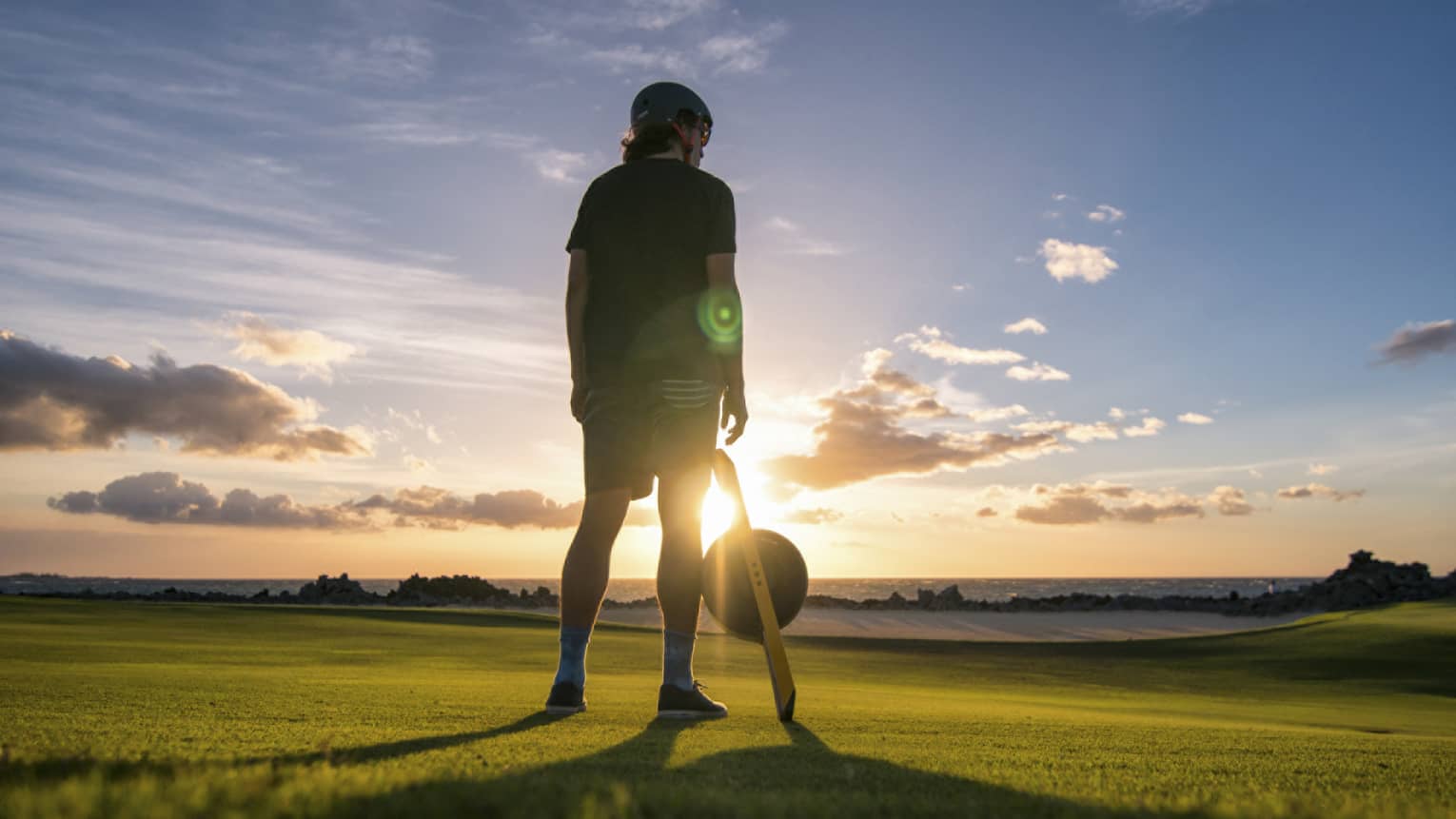 A young boy standing on a large field of green grass with the sun setting in the distance in front of him.