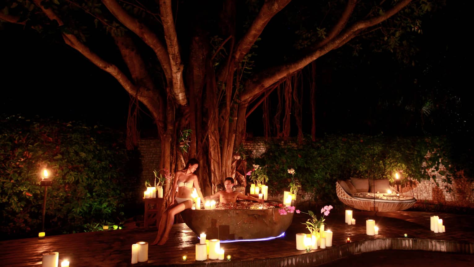 Man in outdoor whirlpool tub at night, woman in bikini on edge, surrounded by lit candles