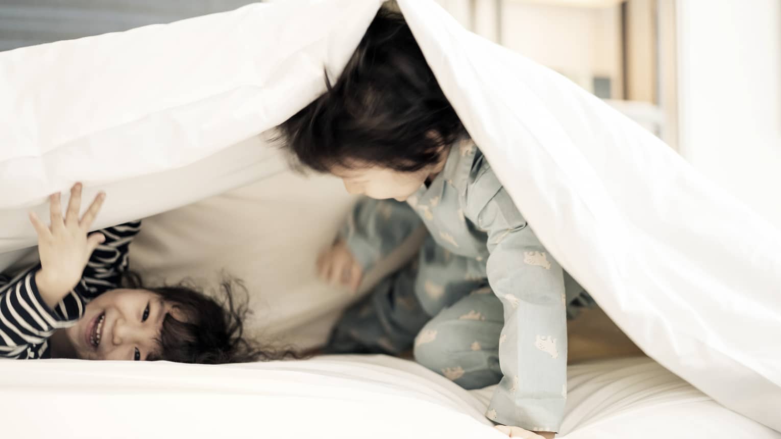 Two young children in pyjamas laugh, play under white blanket on bed