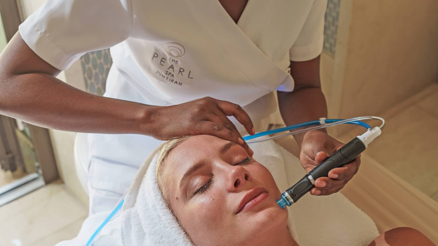 Spa staff holds a blue and black facial tool over woman's face as she lays on massage bed 