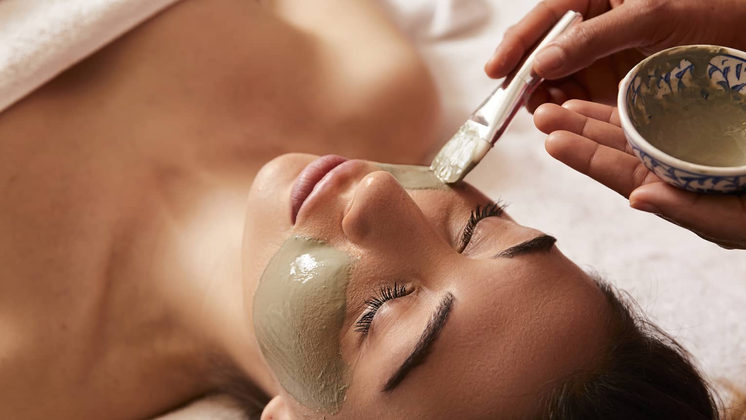 Spa attendant paints clay mask onto woman's face with brush in spa treatment room