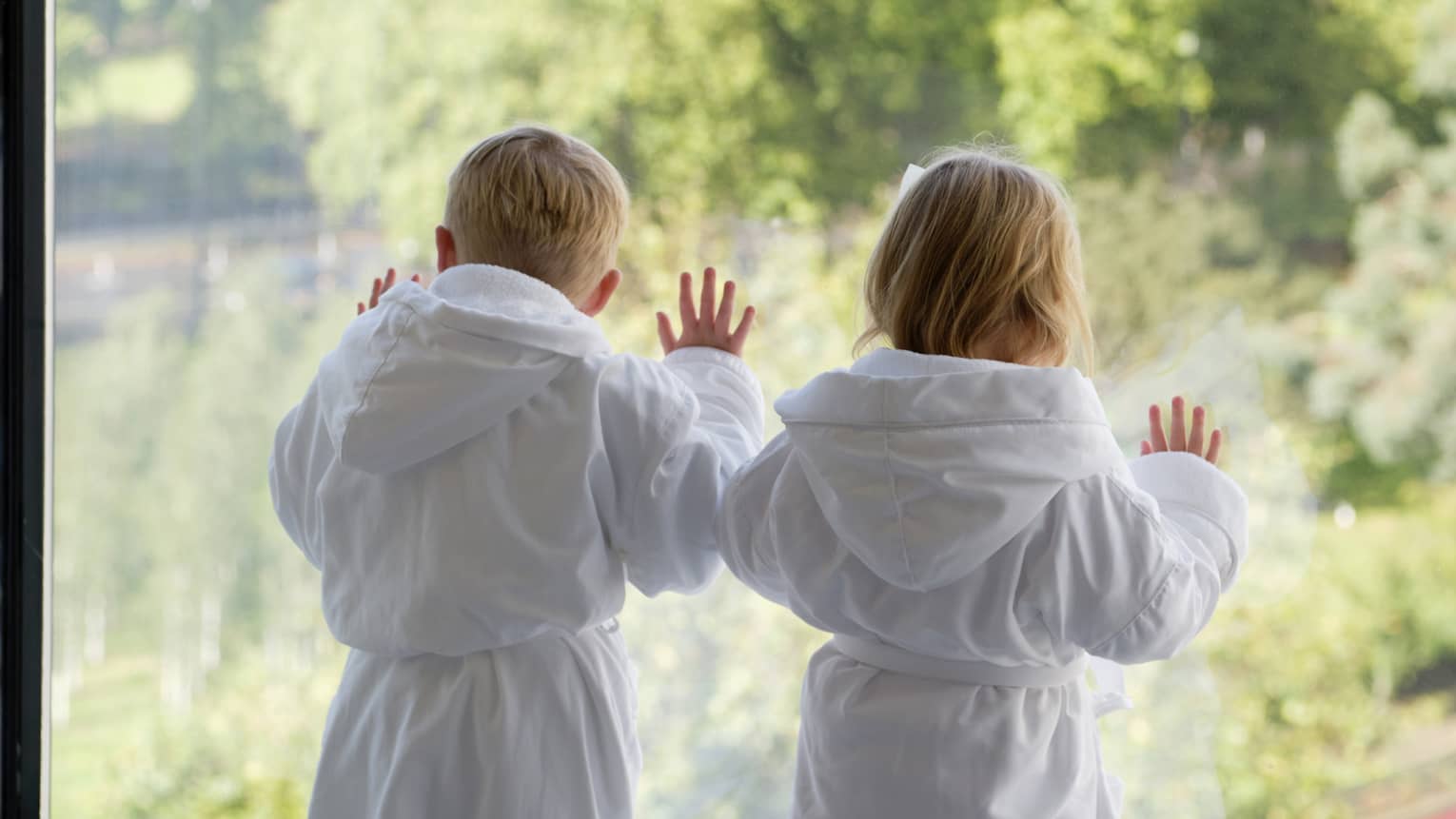 Two young children wearing white bathrobes gaze out floor-to-ceiling window overlooking trees