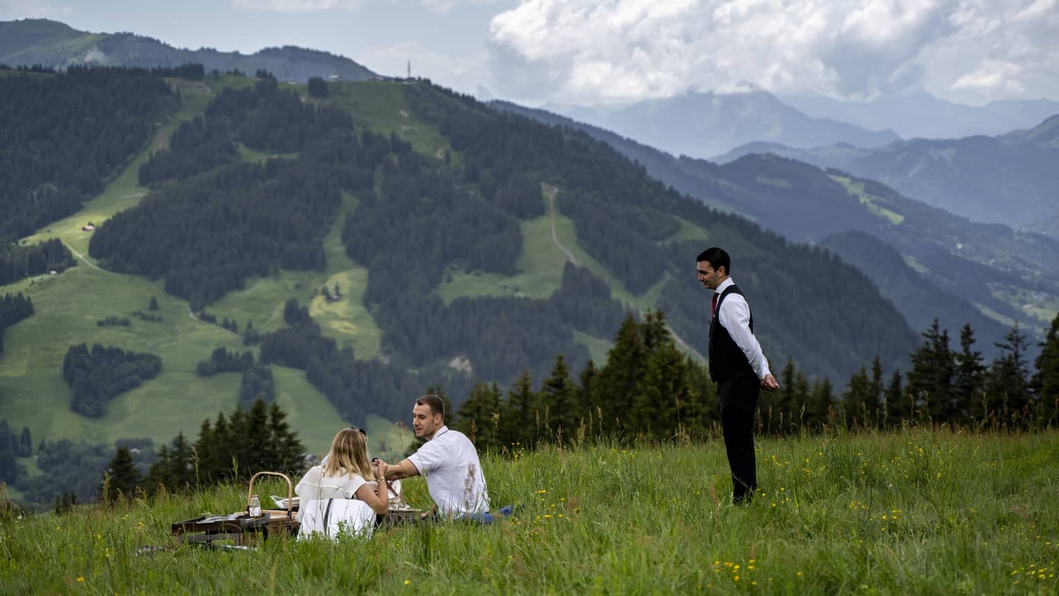 Man and woman sit on blanket with picnic on lush green mountainside, server assisting