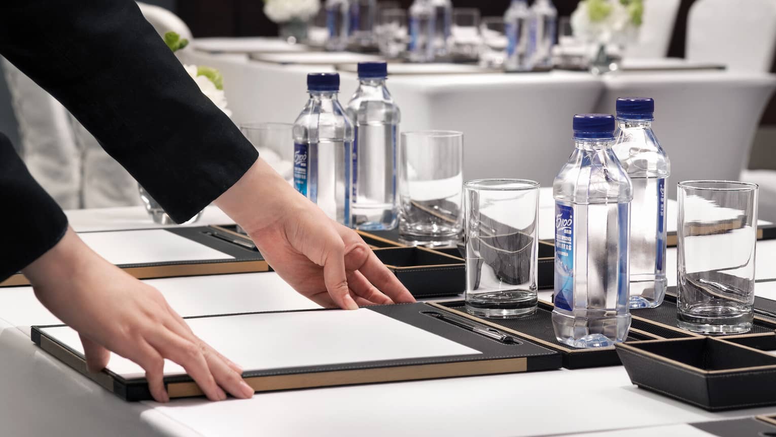 A person adjusts a writing pad on a conference room table with bottled water, glasses and floral arrangements