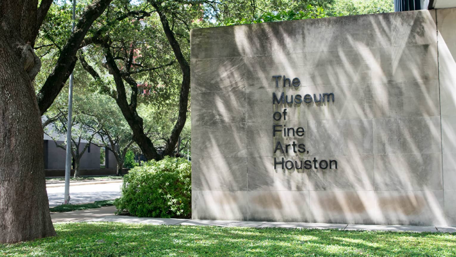 The Museum of Fine Arts Houston sign on stone wall by green lawn, trees