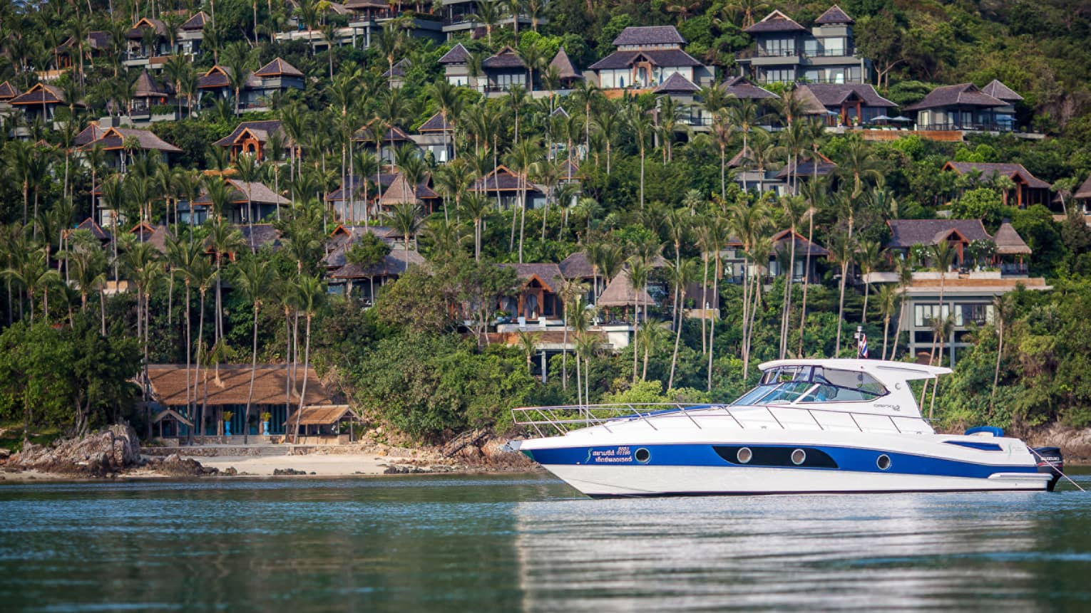 White and blue private fishing boat on ocean in front of mountain dotted with resort villas