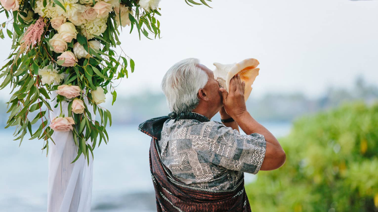 Man holds large seashell to lips at outdoor wedding ceremony