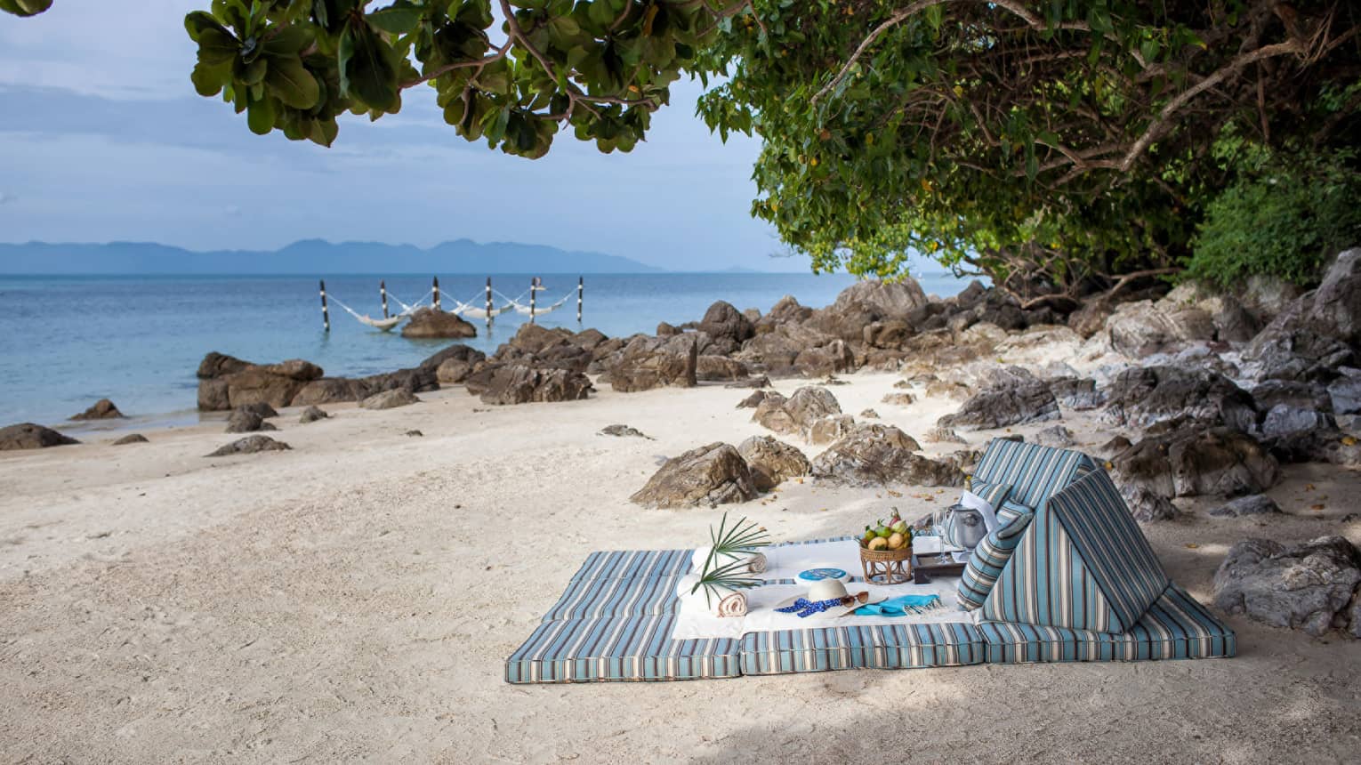 Striped cushions on white sand beach under tree with hat, fresh fruit, towels