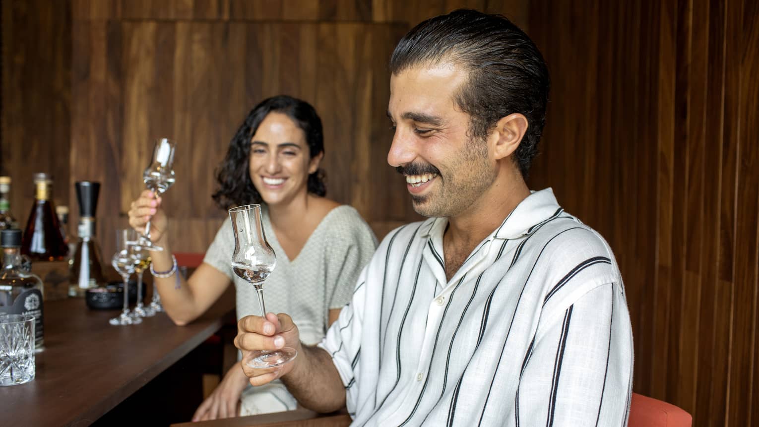 Guests smile while holding grappa glasses filled with agave spirits at a wood bar counter in a wood-panelled room.