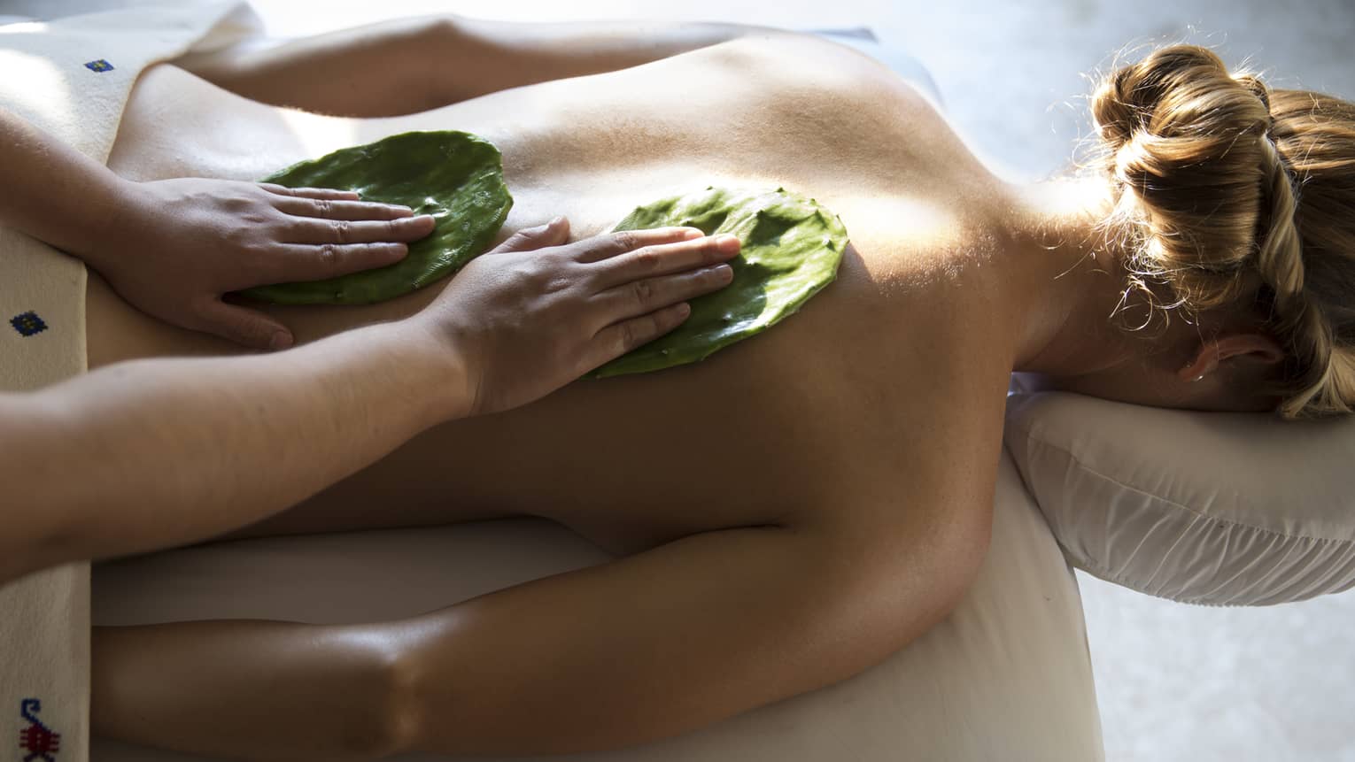 Woman face-down on table as masseuse rubs Nopal cactus leaves on her back with Mexican Nakieri Treatment