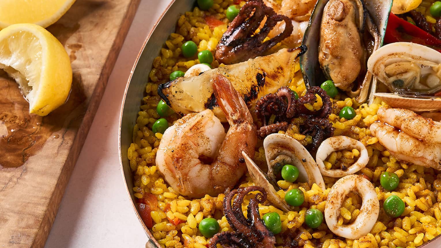 Seafood paella with assorted seafood in traditional oval-shaped metal pan, lemon wedges on cutting block
