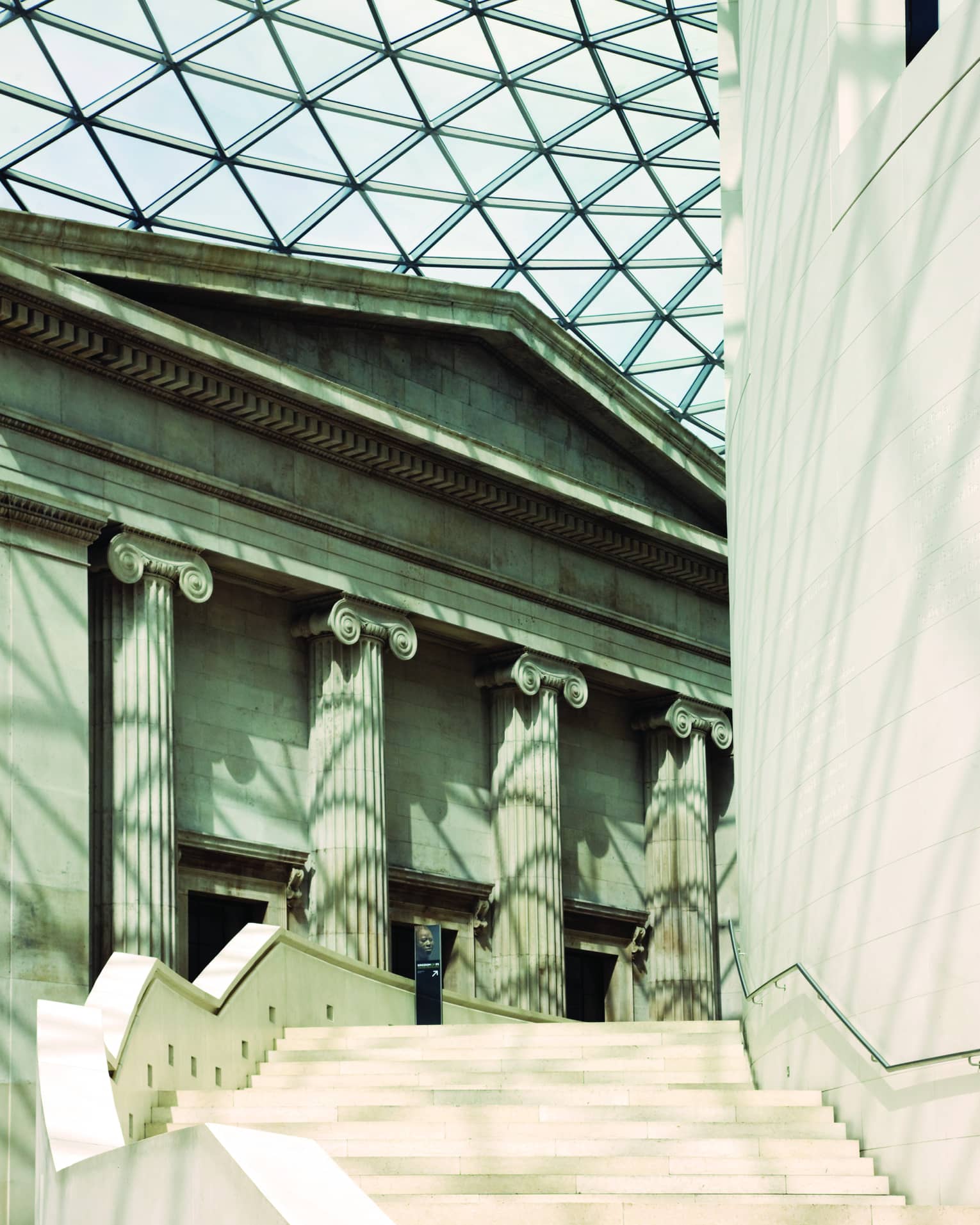 Close-up of façade and staircase in the British Museum courtyard, light through the glass roof leaving criss-cross shadows.