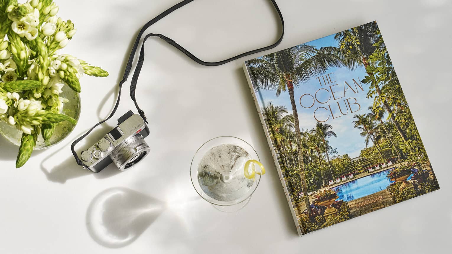 A large book with a pool and palm trees on the cover next to a drink and camera.