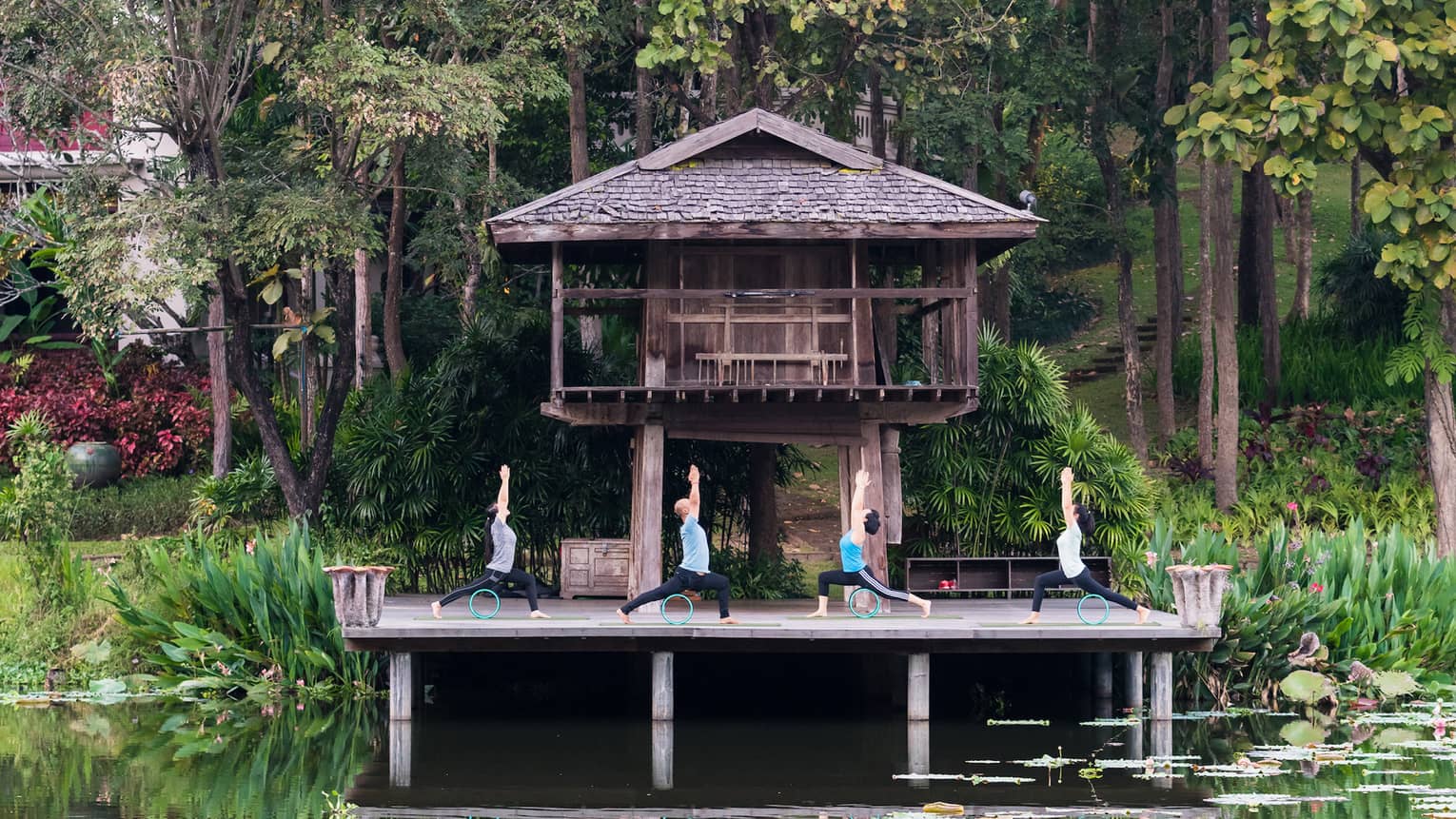 Four people doing yoga on a deck overlooking a calm pond, in front of a wooden bungalow and jungle trees