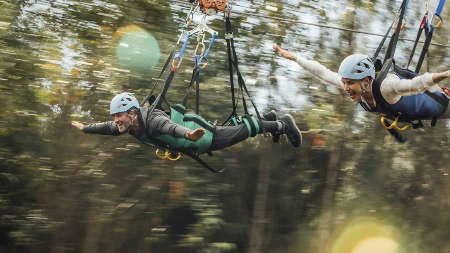 Close-up of a couple in full safety gear with wide smiles and arms outstretched as they zip-line past a blur of treetops.