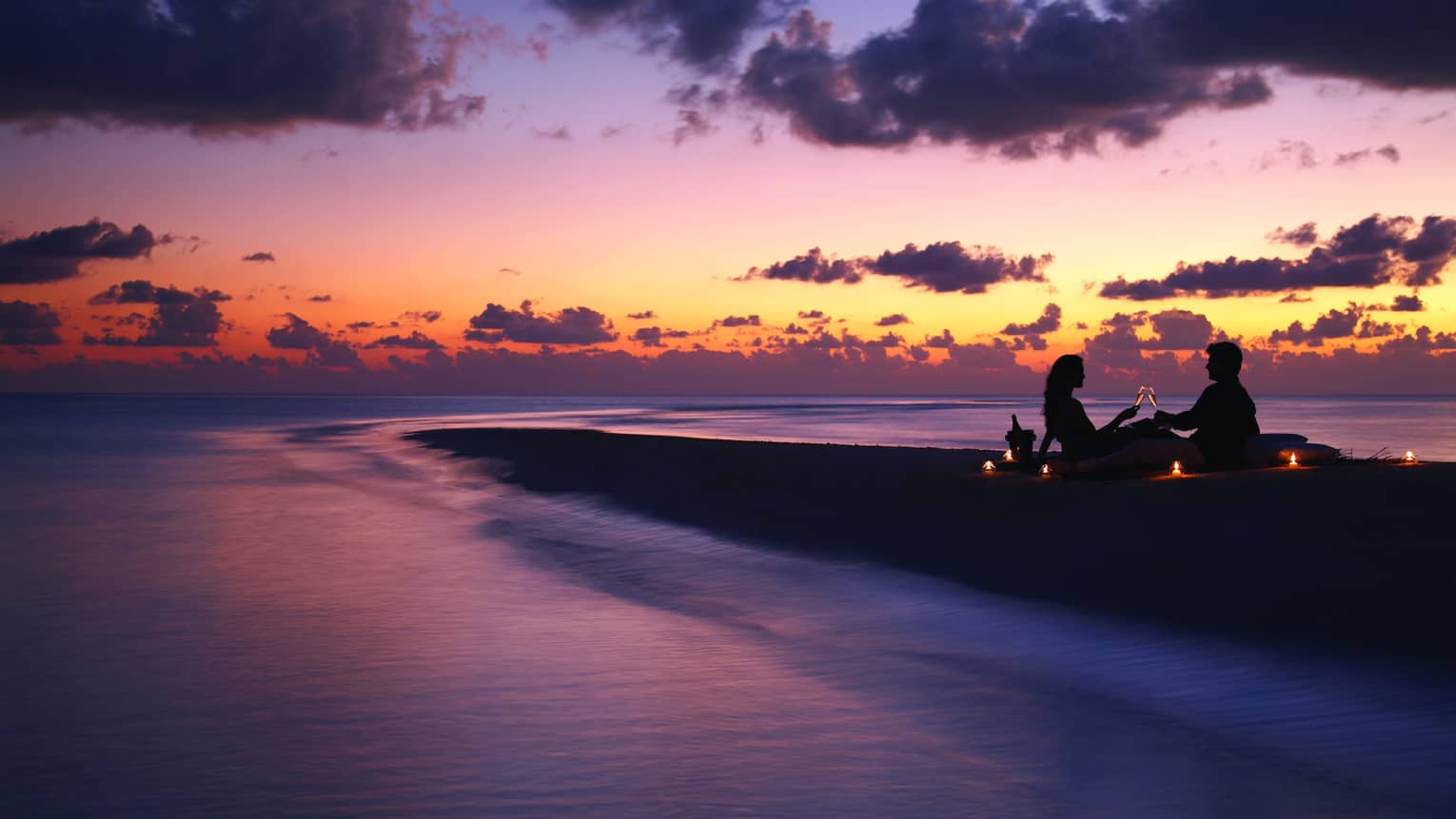 Sandbank dinner, silhouette of couple sitting beach, toasting Champagne at sunset