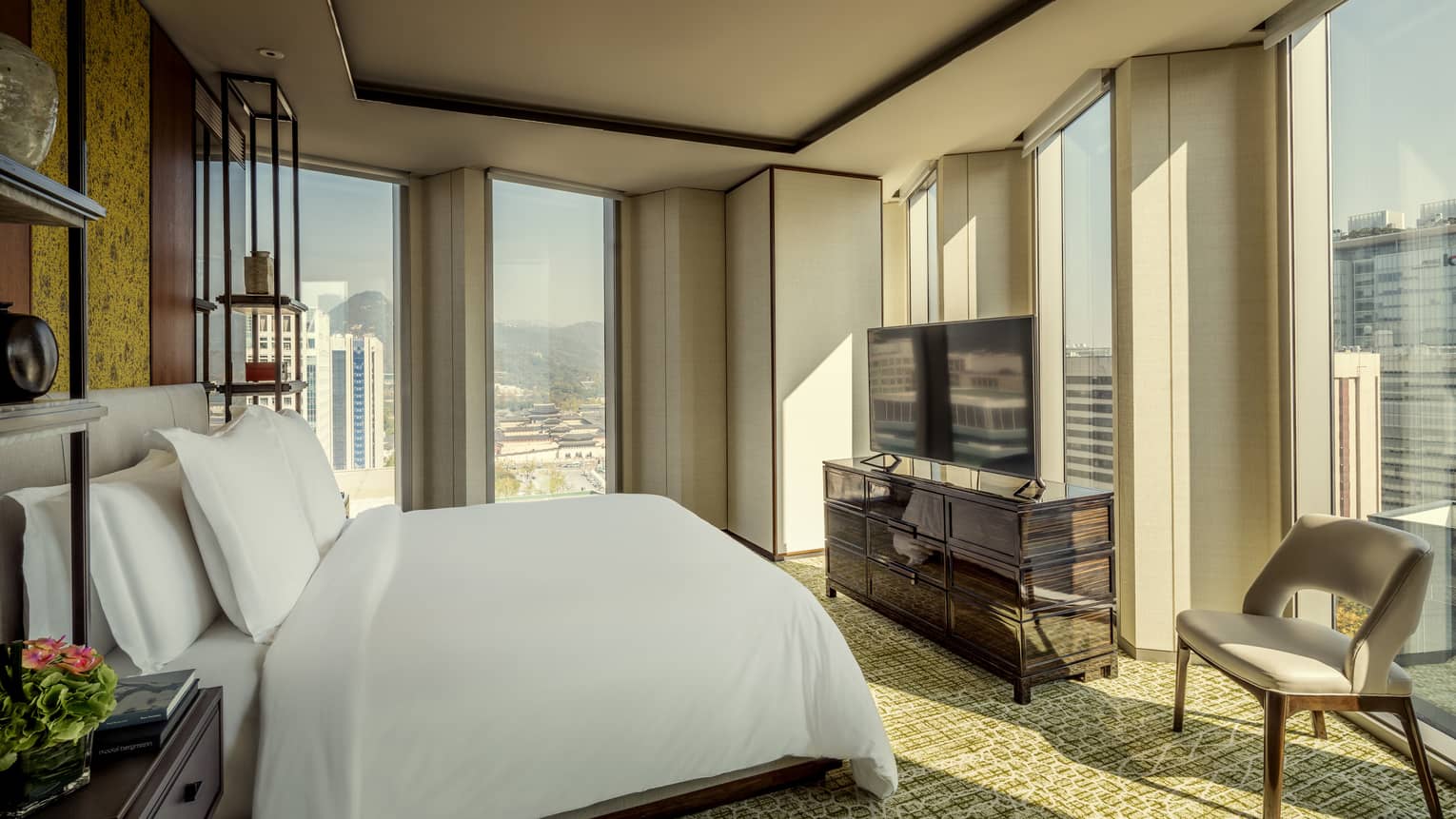 Corner guest room with bed and white linens, dresser and flatscreen TV, armchair, floor-to-ceiling windows with city views