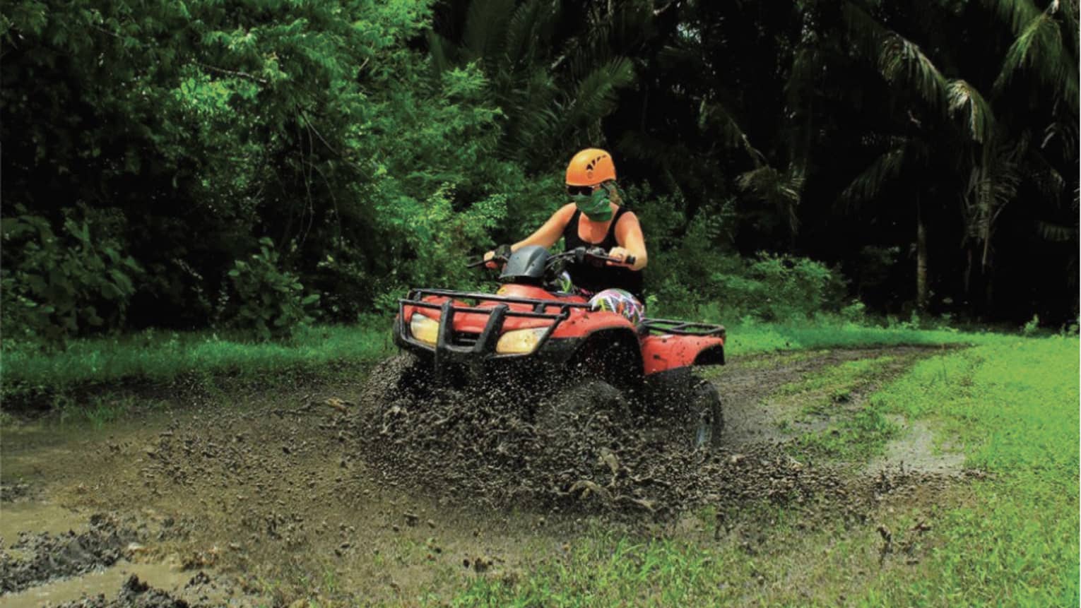 Person wearing a black tank top and bright orange helmet rides through the mud on a red ATV