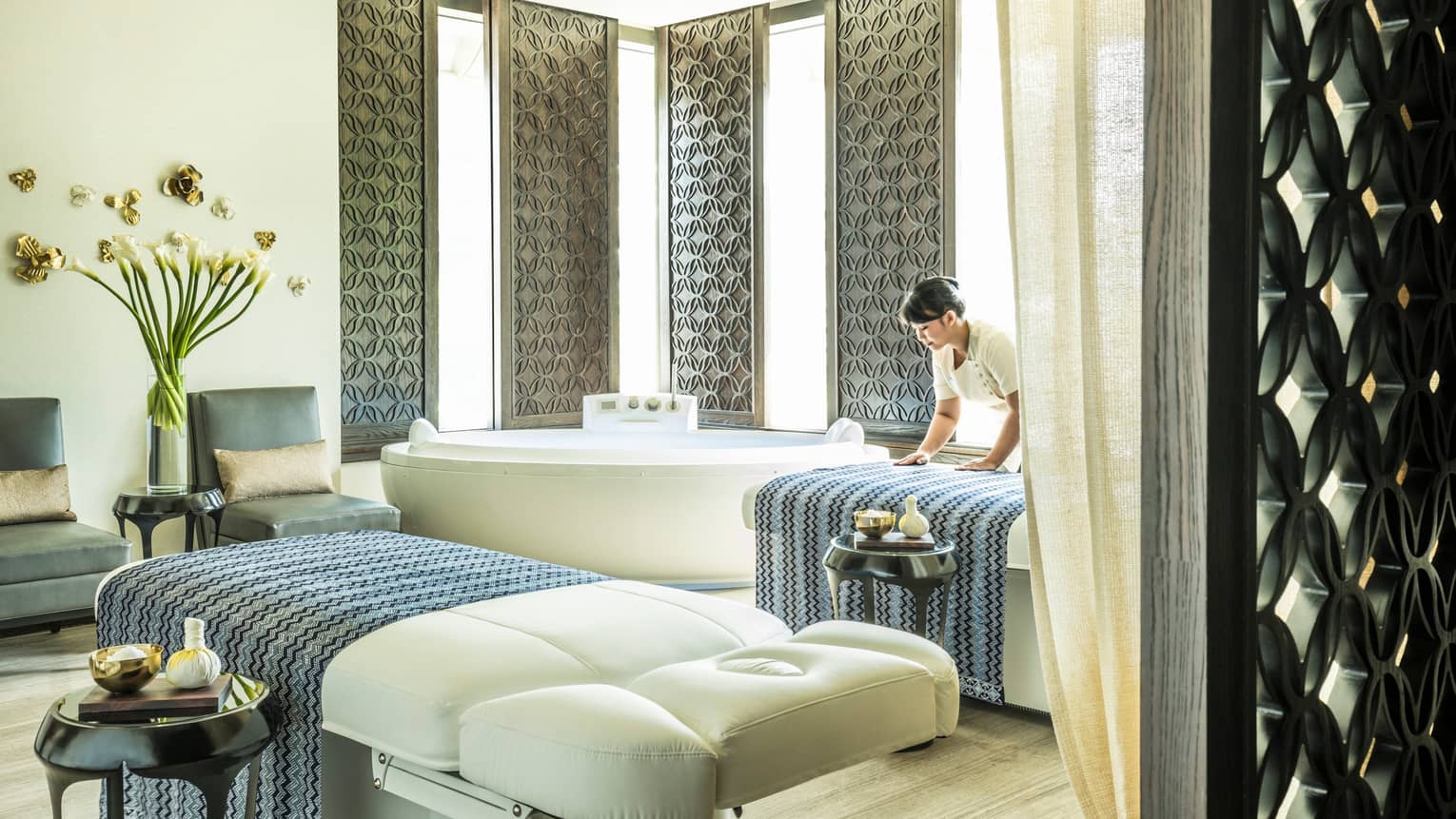 Large white spa table in front of white spa tub, silver wall panels, woman places blanket on table