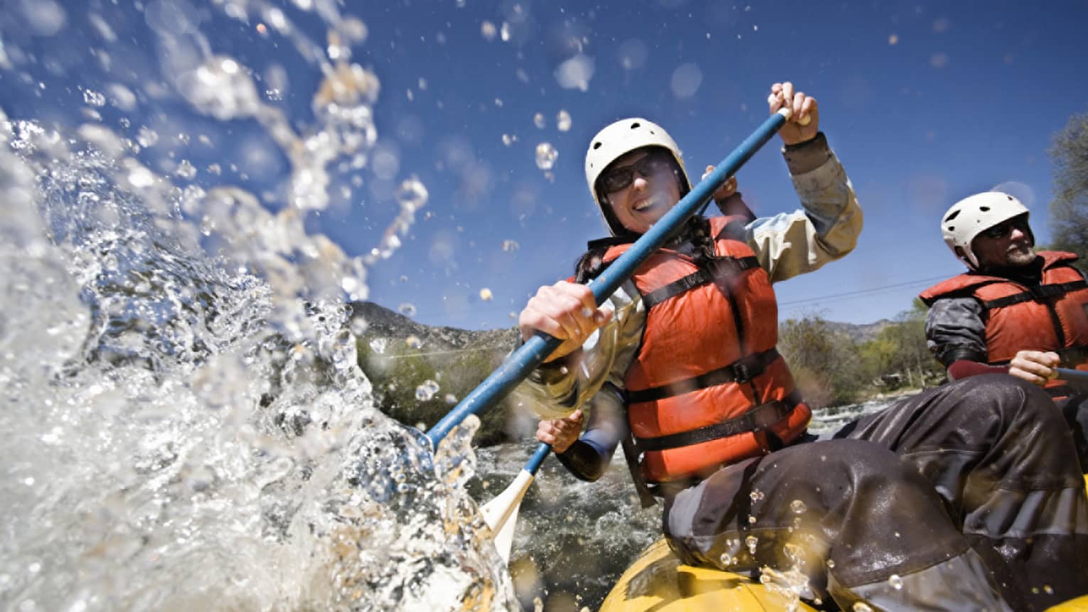 Smiling woman wearing red life vest holds paddles as waves crash over raft