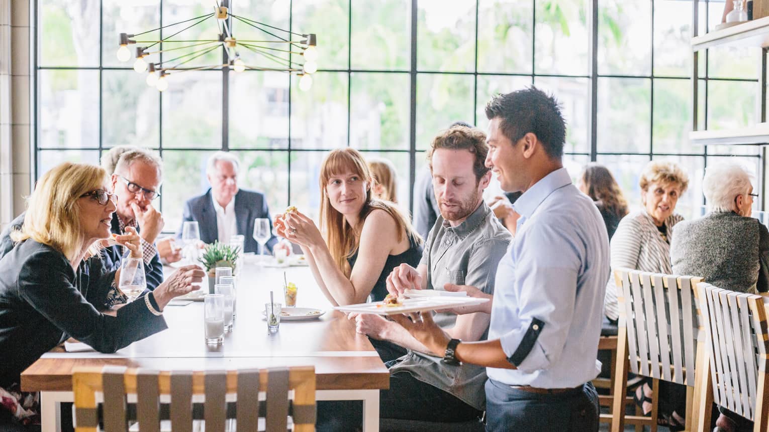 Crowds gathered around long dining table in sunny Seasons Restaurant as server stands with tray of appetizers