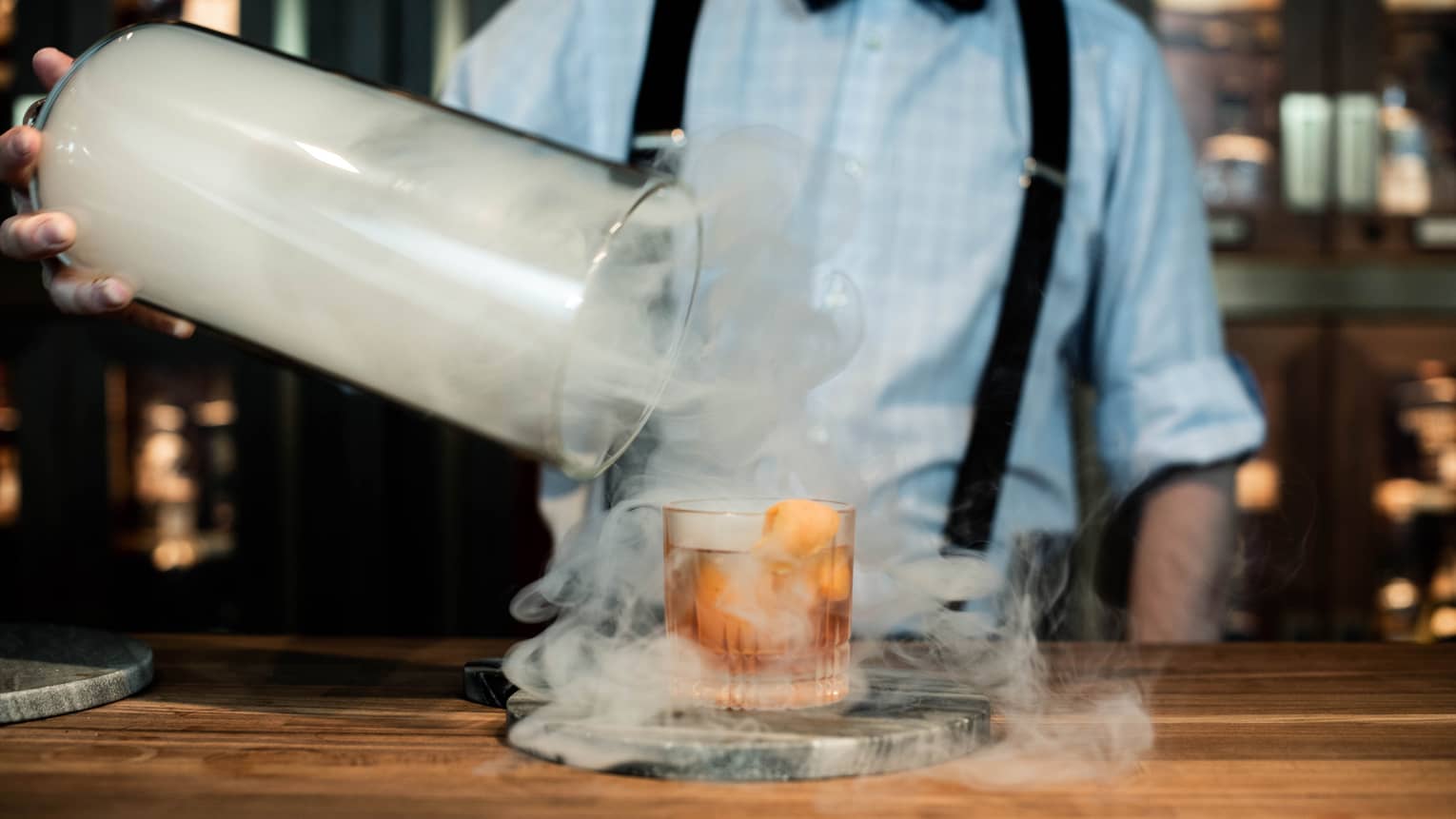 Smoke swirls around a cocktail as a bartender removes a lid 