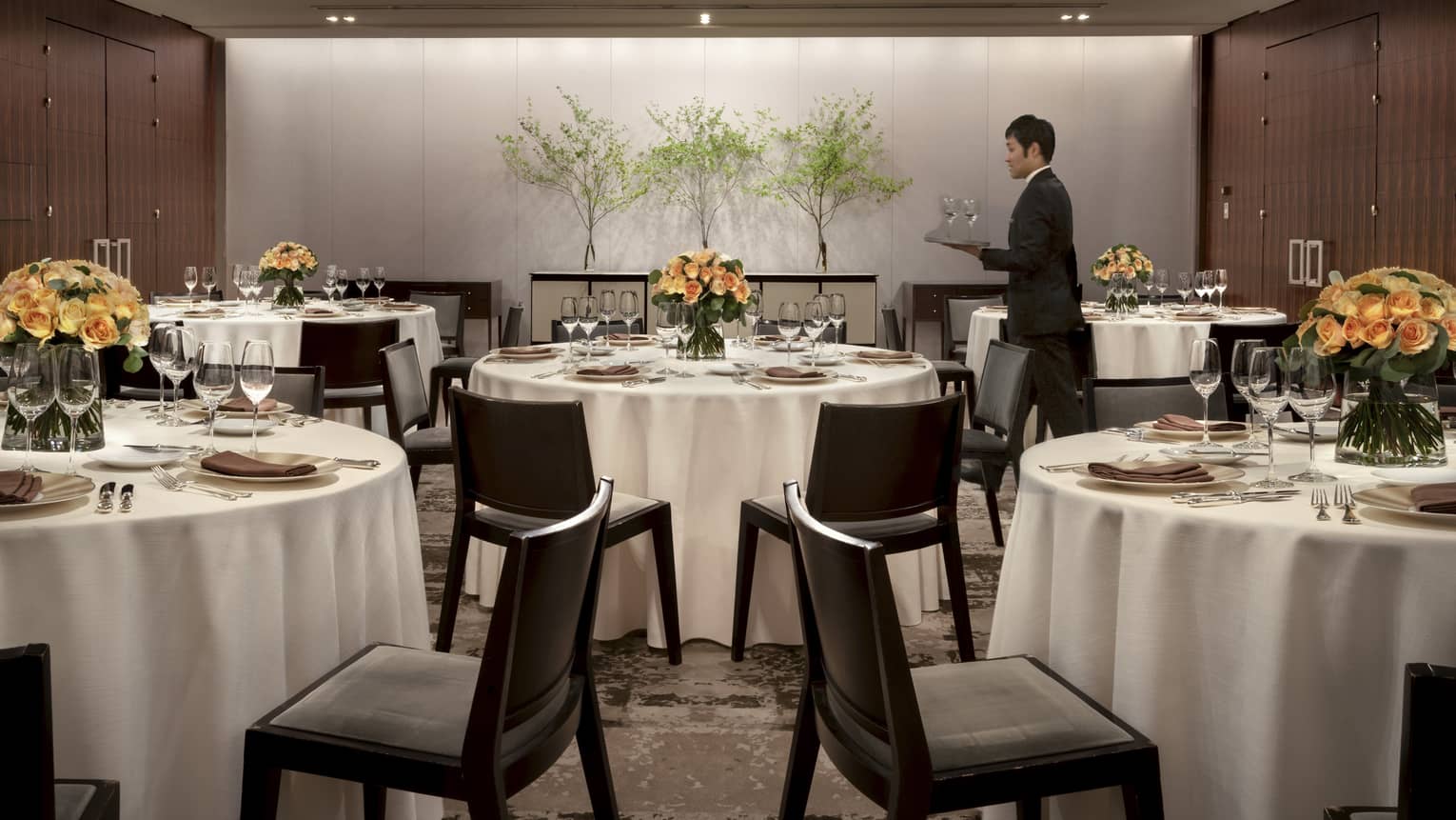 Hotel waitstaff carries tray with wine glasses past small round banquet tables