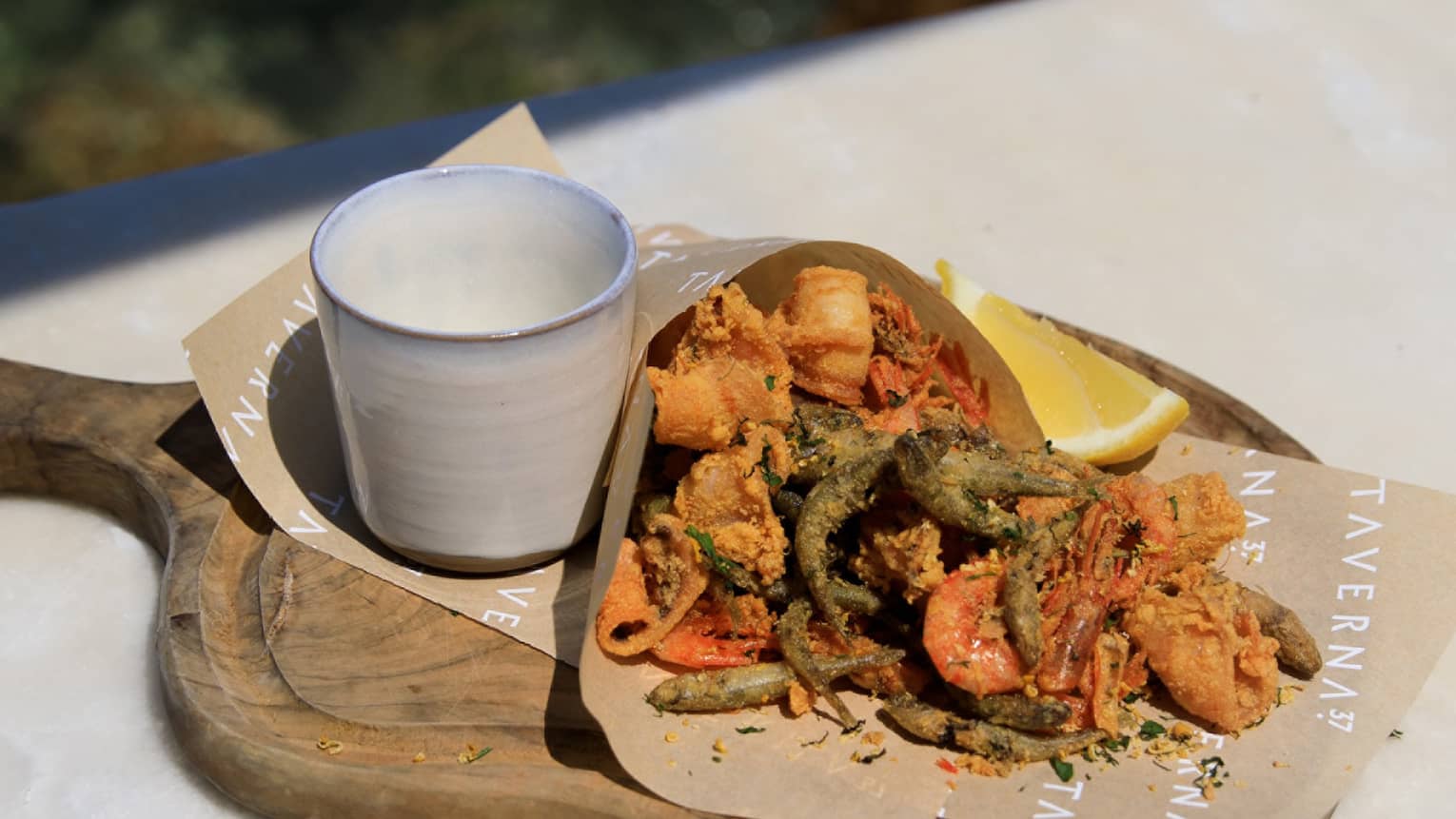 Fried seafood wrapped in paper on cutting board with cup of caper sauce and lemon wedge