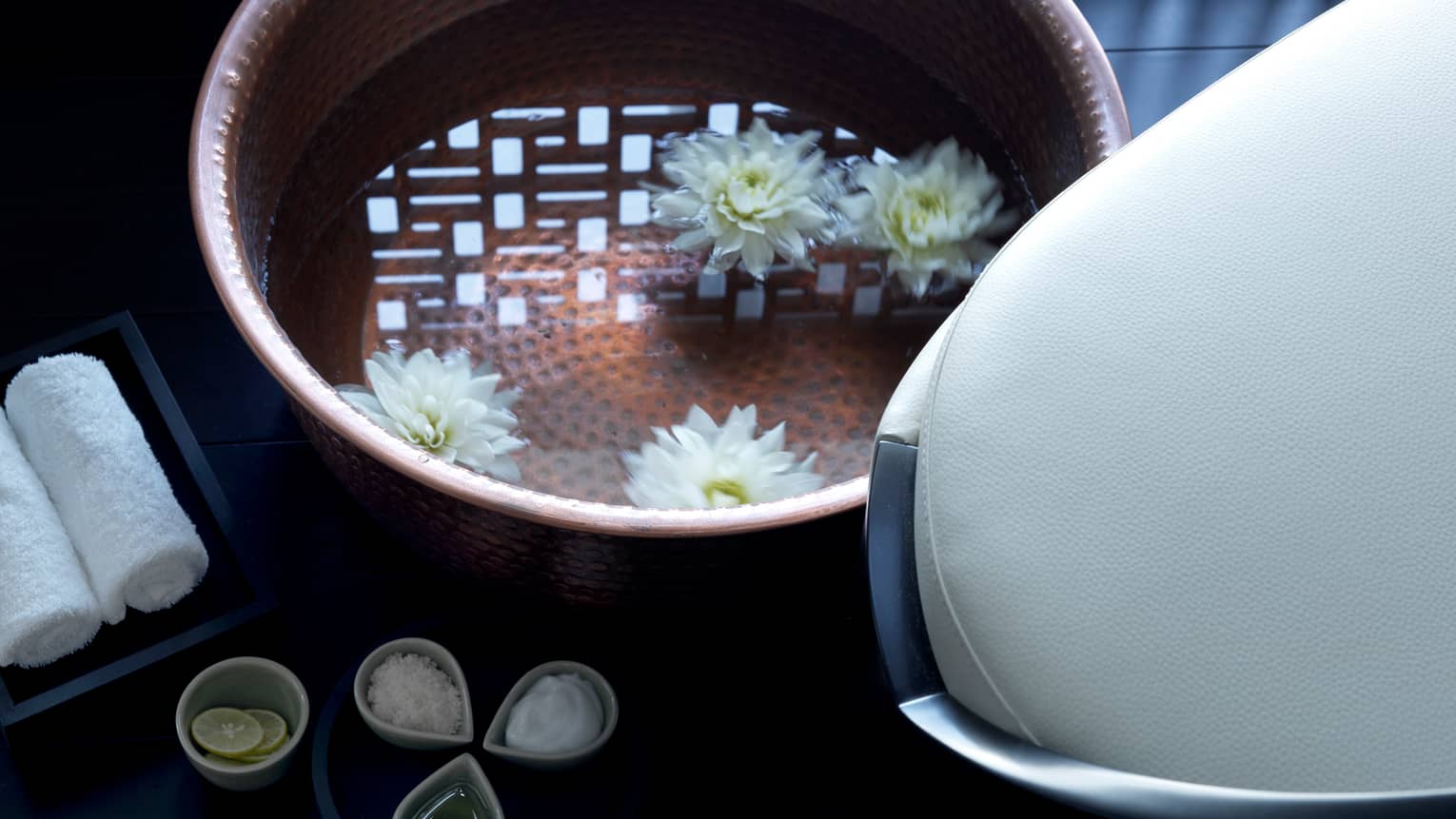 Close-up of copper bowl with water, floating white flowers, small dishes with spa salts, oil and lemons