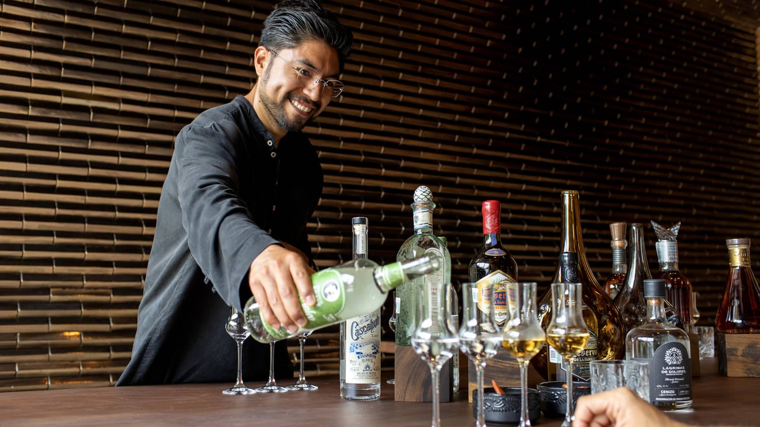 A bartender pours a clear liquor into grappa glasses, two others filled with an amber liquor, aside various other bottles.