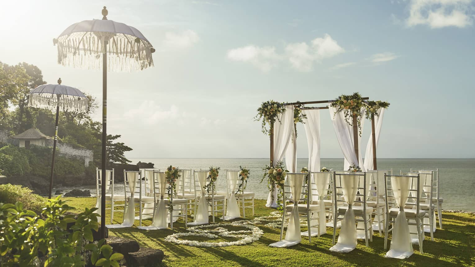 Sun shines over wedding altar draped in white fabric, rows of chairs overlooking ocean