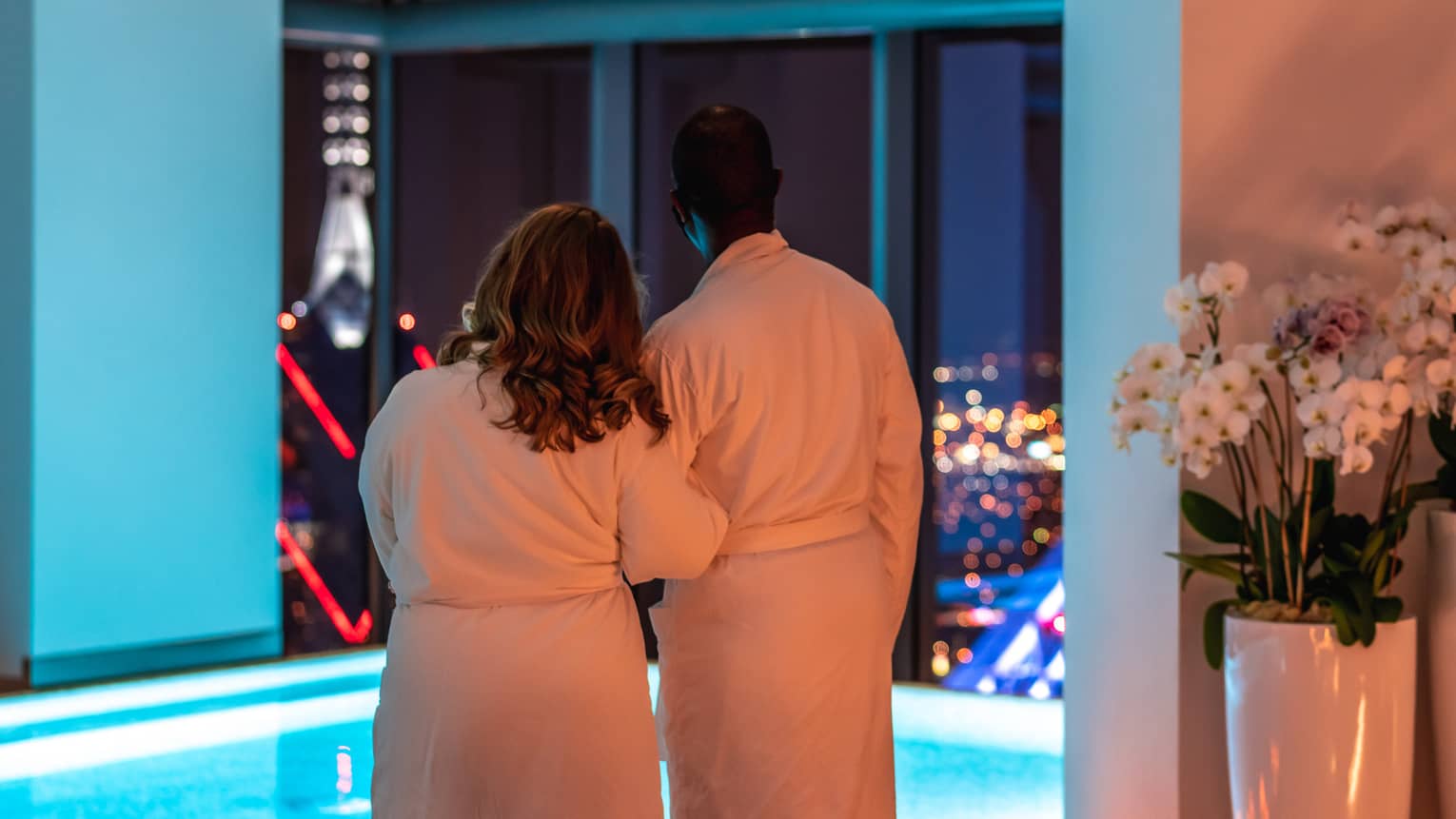 A man and woman in spa robes looking out a large window at a city during night.